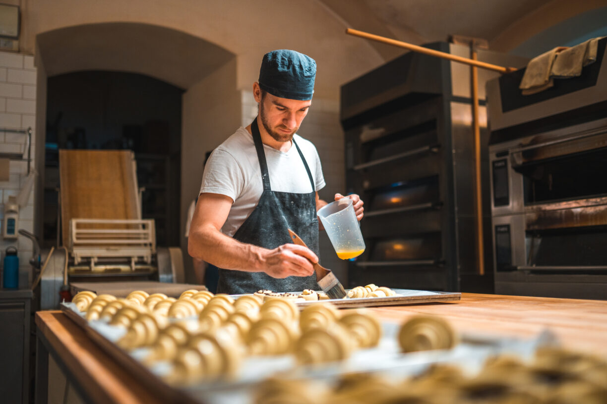 baker applying egg wash on to raw pastries on a baking trays before going into an oven